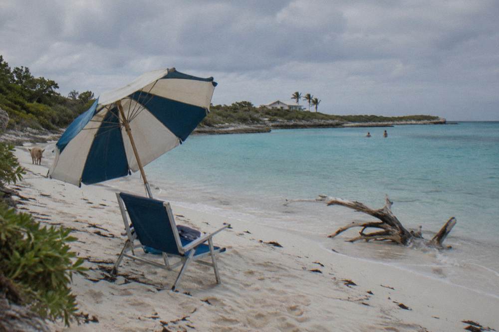 A shaded beach chair and umbrella on white sand facing calm blue water