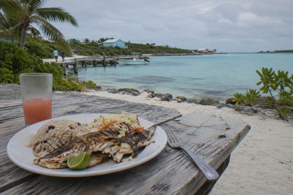 A wooden table with a Bahamian lunch of grilled fish and sides near a beach