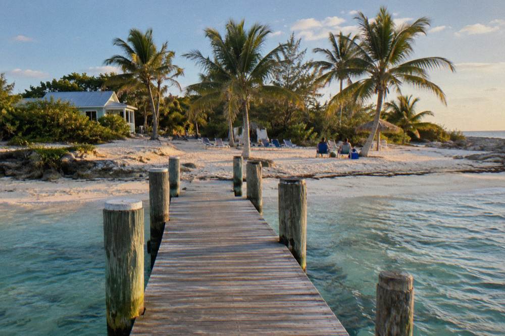 Wooden dock extending over clear shallow water toward a small beach with beach chairs