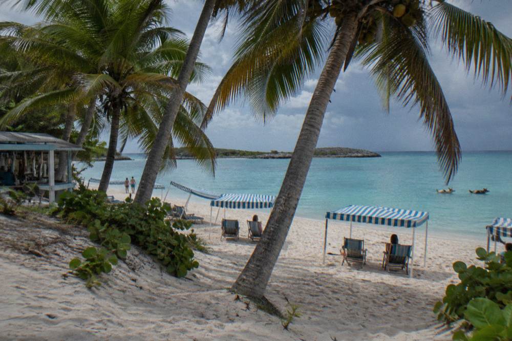 Shaded beach chairs and palm fronds on a small island beach beside calm pale blue water