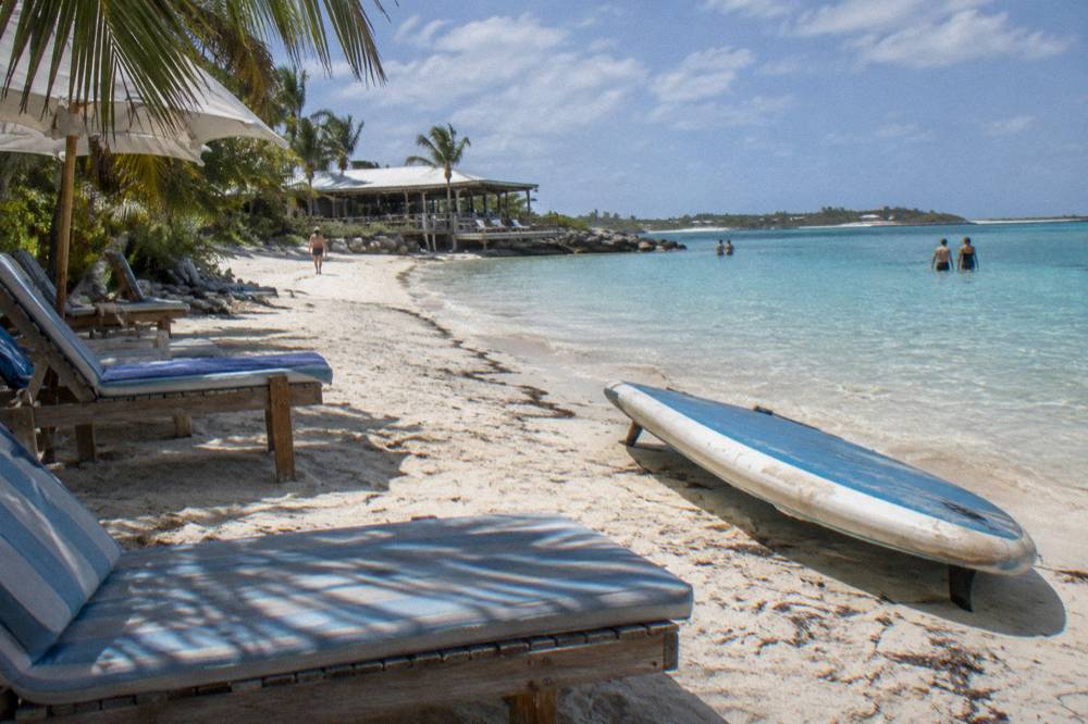 Shaded beach loungers and a paddleboard on sand beside calm shallow water