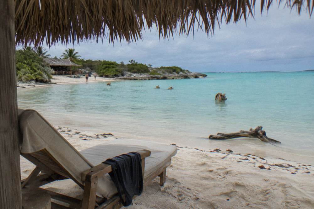 A shaded beach chair and umbrella on white sand beside calm shallow turquoise water